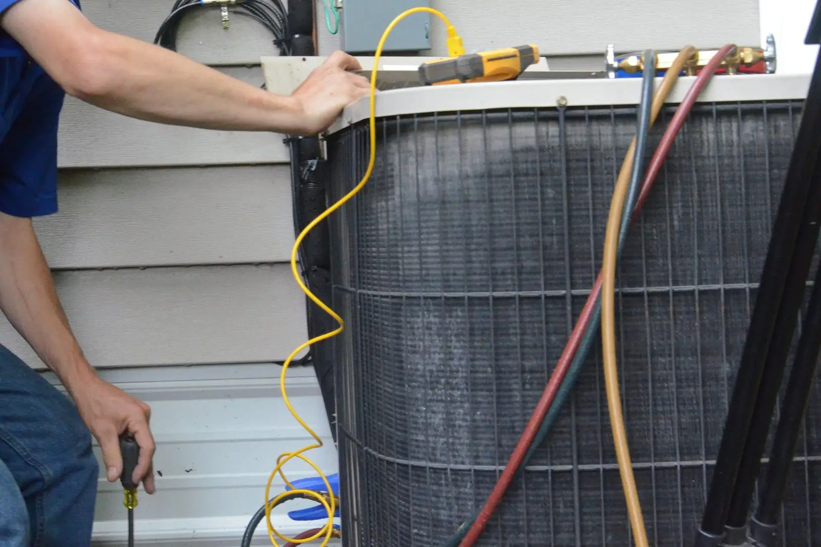 Technician checking an outdoor AC condenser with a clamp meter and refrigerant hoses