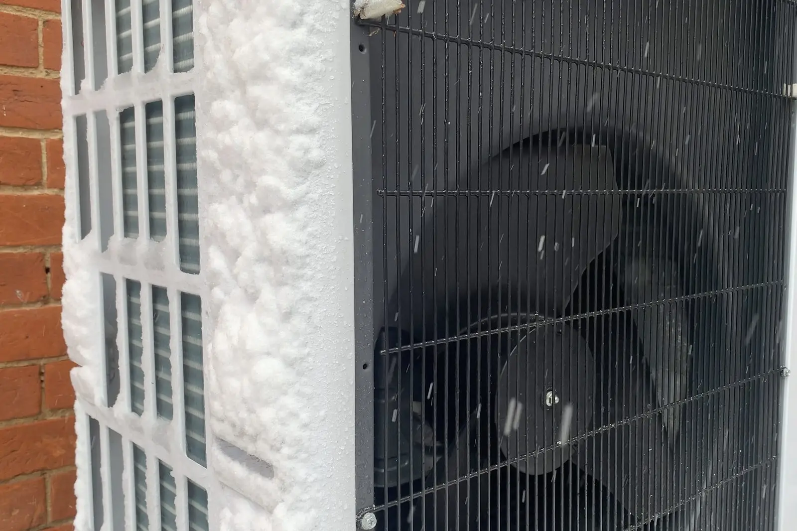 Snow-caked heat pump outdoor unit running against a brick wall during snowfall